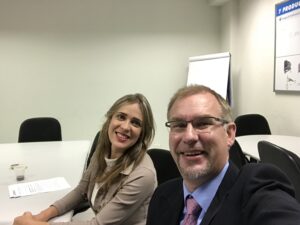 Inglés profesional: Steven y Gisele sentados a una mesa de conferencias. Él está a la derecha, sonriendo, con gafas, traje oscuro y camisa azul. Junto a él, Gisele sonríe; tiene el cabello largo y rubio y viste una chaqueta beige.