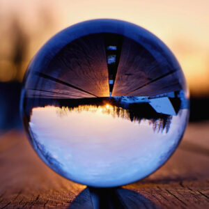 A crystal ball, on a wooden surface, with an upside-down reflection of snow, trees, and a house under a warm sunset