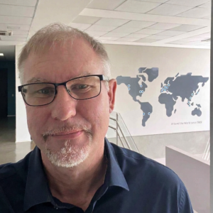 Selfi of Steven in a dark blue shirt. In the background, a grey map, with the words "Around the World since 1969" below.