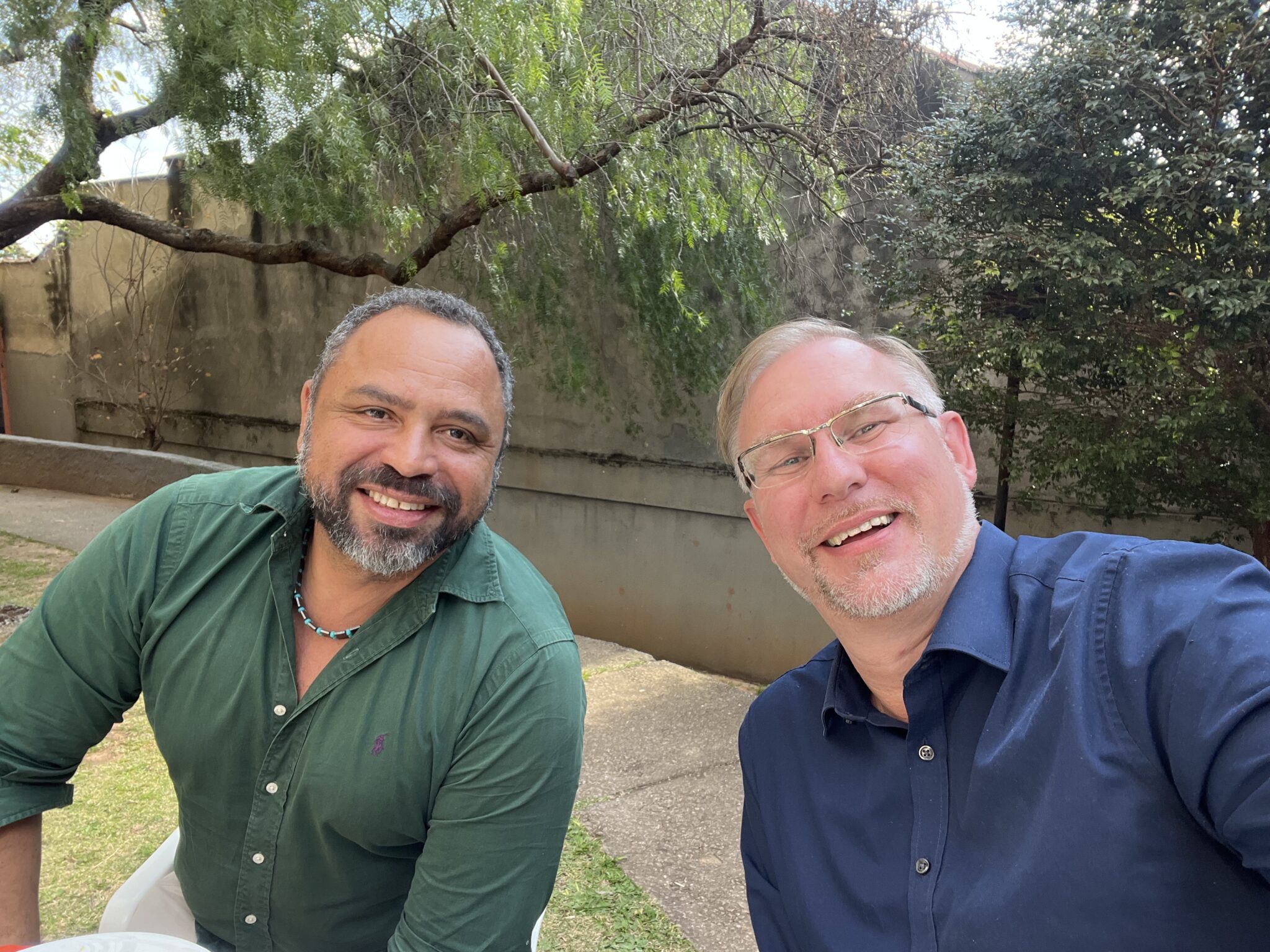 Ari and Steven in an informal outdoors garden setting, Ari left, dark green open collard shirt, Steven right, dark blue open collared shirt, both relaxed and smiling.